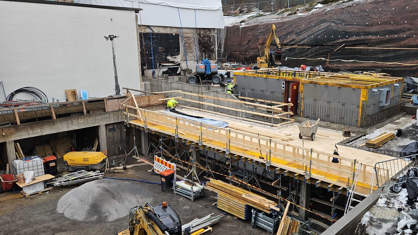 Rakennustyömaalla näkyy betonirunkoja ja -pilareita sekä työmaakoneita. Taustalla vanhan museorakennuksen valkoiseksi maalattu tiiliseinä sekä louhittu kallioseinä.